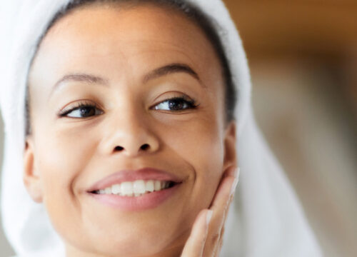 Photo of a smiling woman touching her face. Her hair is wrapped in a towel after a shower.
