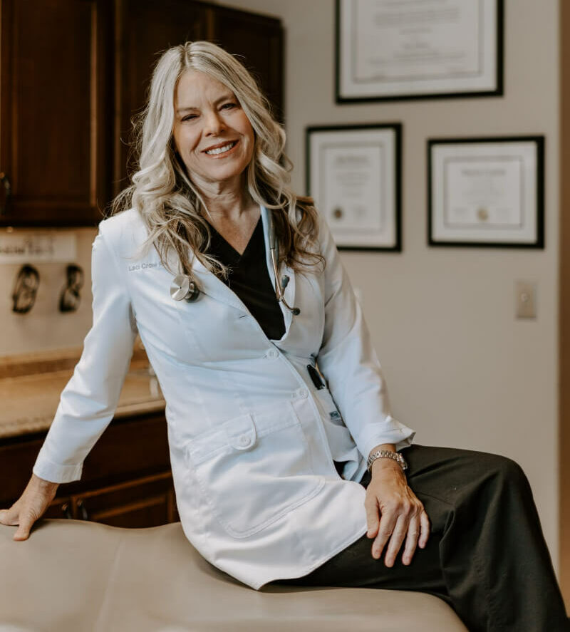 A woman in a white lab coat sits on an exam table in a medical office with framed certificates on the wall behind her.