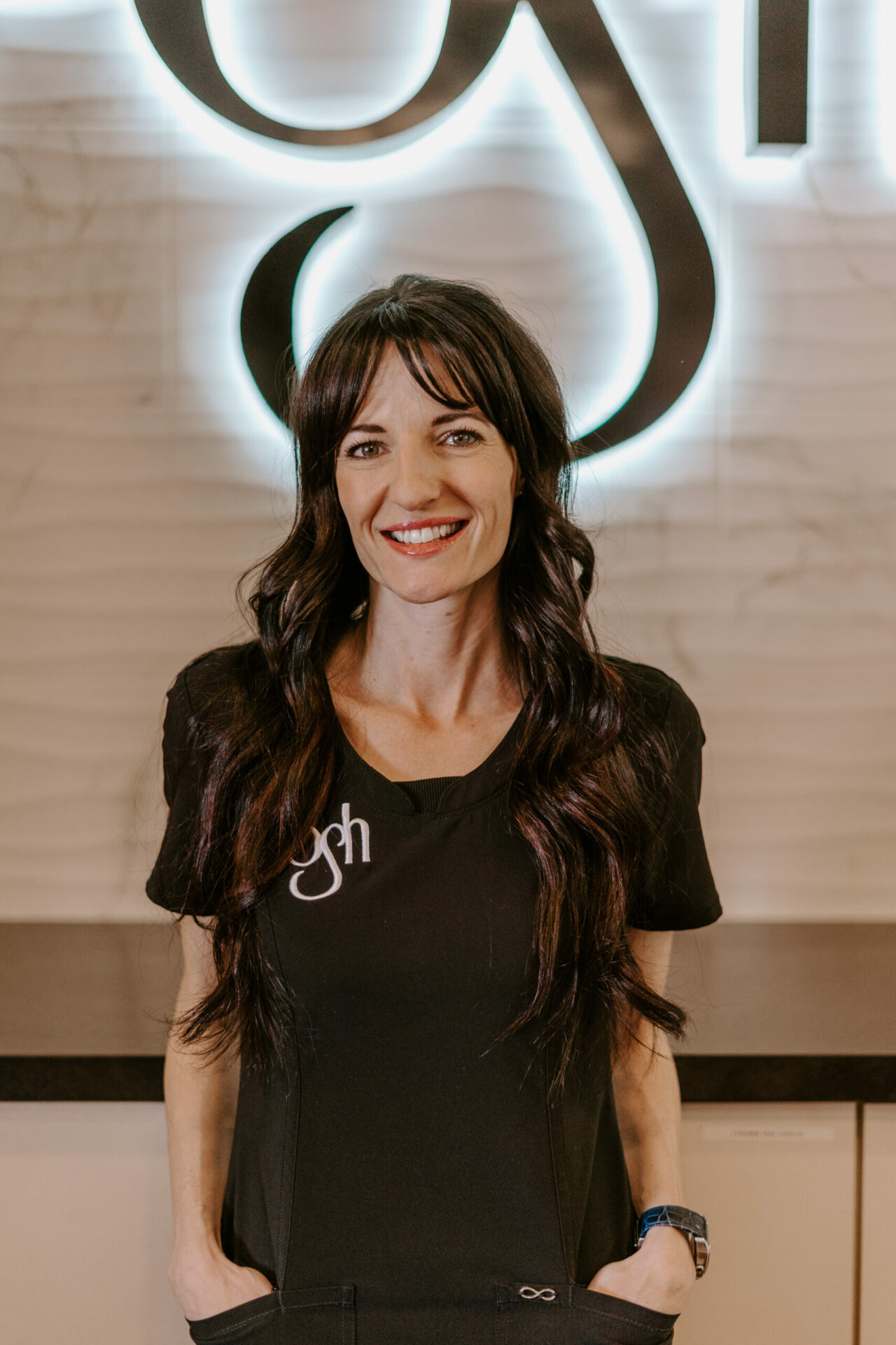 Ayla Elizabeth Pike, a woman with long dark hair and a black top with "gh" embroidered on it, stands smiling in front of a lit wall sign.