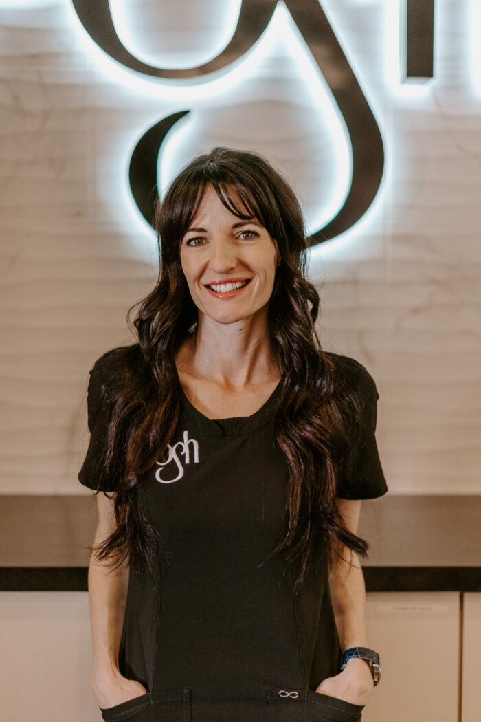 Ayla Elizabeth Pike, a woman with long dark hair and a black top with "gh" embroidered on it, stands smiling in front of a lit wall sign.