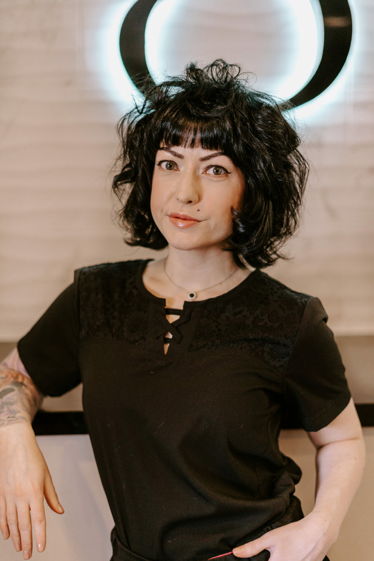 Heather Tolson, a woman with curly dark hair and tattoos on her arm, stands in front of a wall with a circular light glowing behind her head.