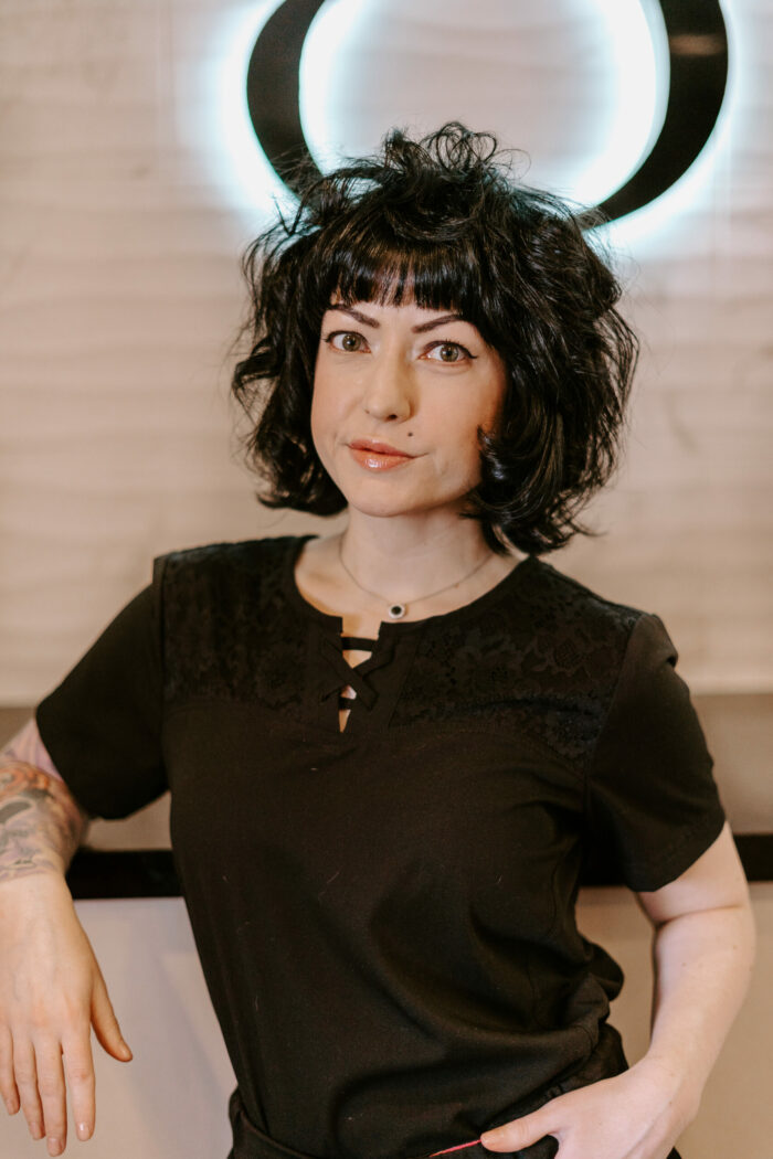 Heather Tolson, a woman with curly dark hair and tattoos on her arm, stands in front of a wall with a circular light glowing behind her head.