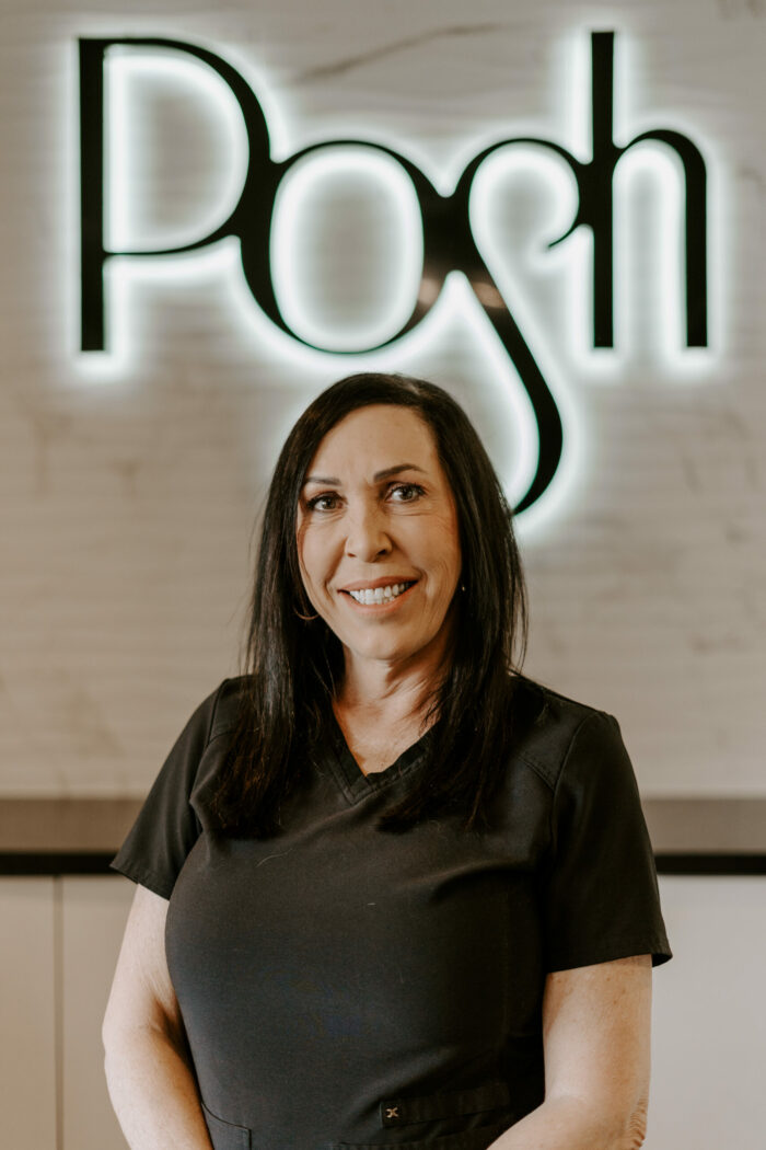Suzette Bennet, with long dark hair and a black top, stands in front of a wall illuminated by the large word "Posh.