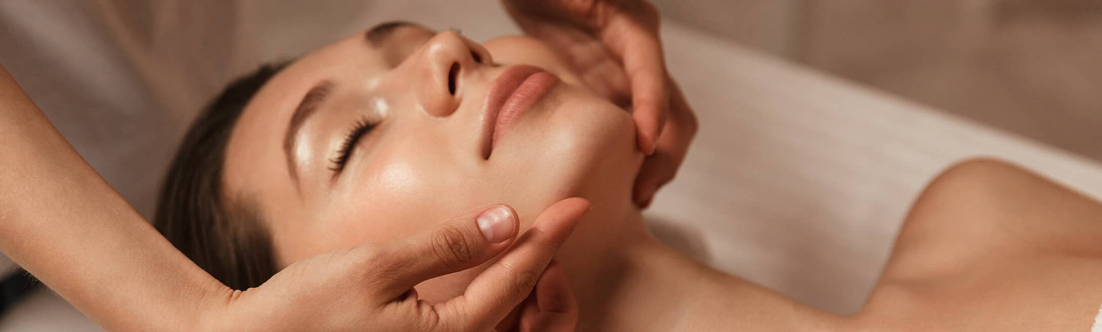 A woman lies with her eyes closed as another person gently massages her face with both hands in a spa-like setting.
