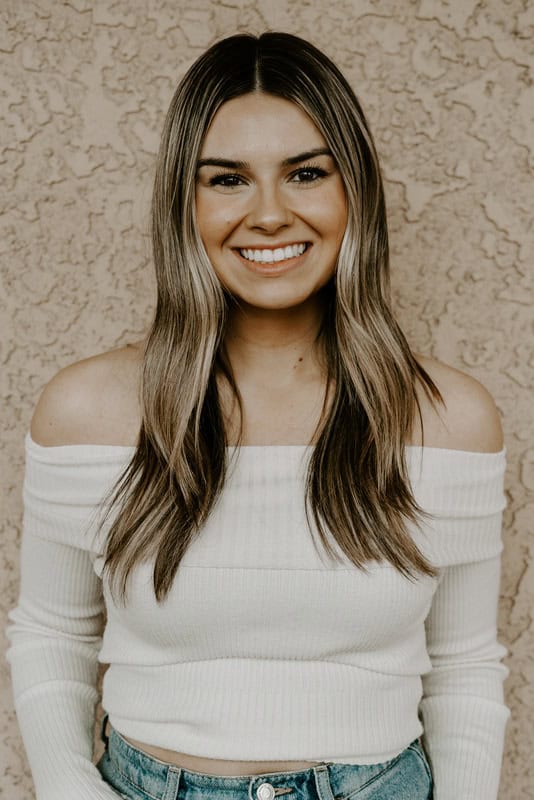 Lauren DiMaggio, with long brown hair, wears an off-the-shoulder white top and blue jeans as she stands against a textured beige wall, smiling at the camera.