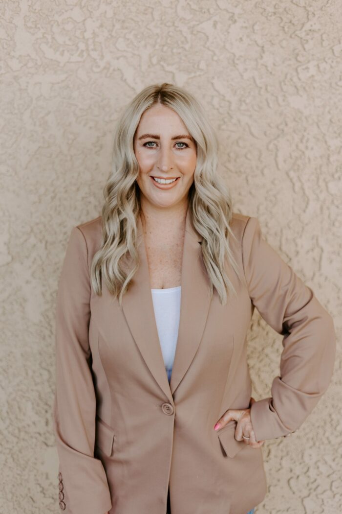 Mary Petrilena, a woman with long blonde hair wearing a beige blazer and white top, stands in front of a textured light-colored wall, smiling at the camera.