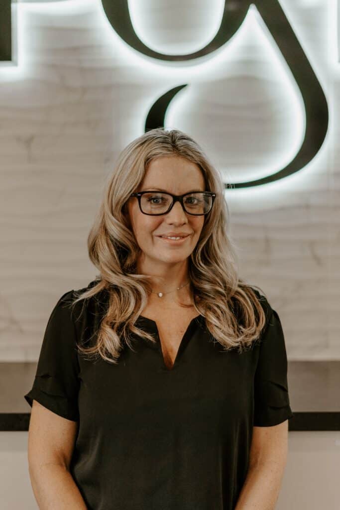 A woman with long blonde hair and glasses stands indoors, wearing a black blouse, in front of a wall with a large illuminated design.