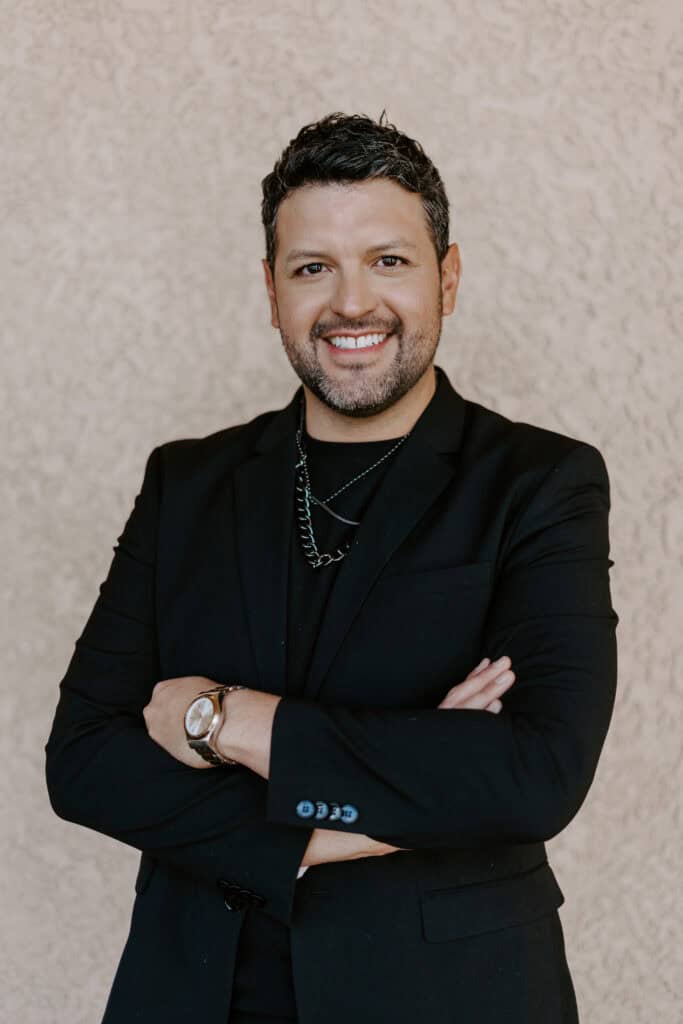 Man with short dark hair and a beard, wearing a black suit, black shirt, and chain necklace, standing with arms crossed and smiling in front of a textured beige background.