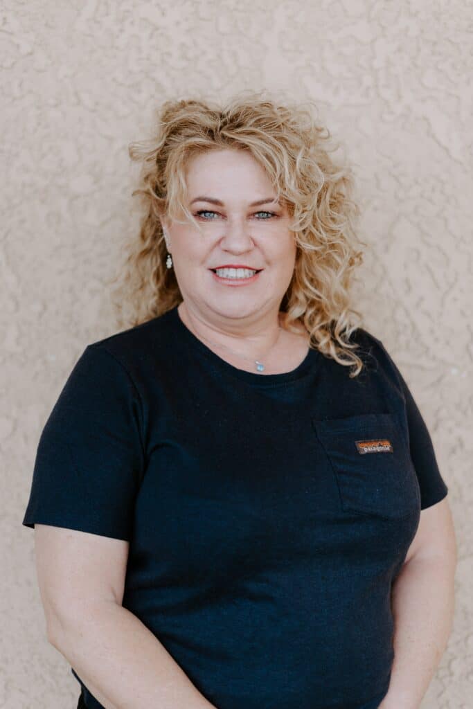 A woman with curly blonde hair, wearing a navy blue t-shirt, stands in front of a textured beige wall, smiling at the camera.