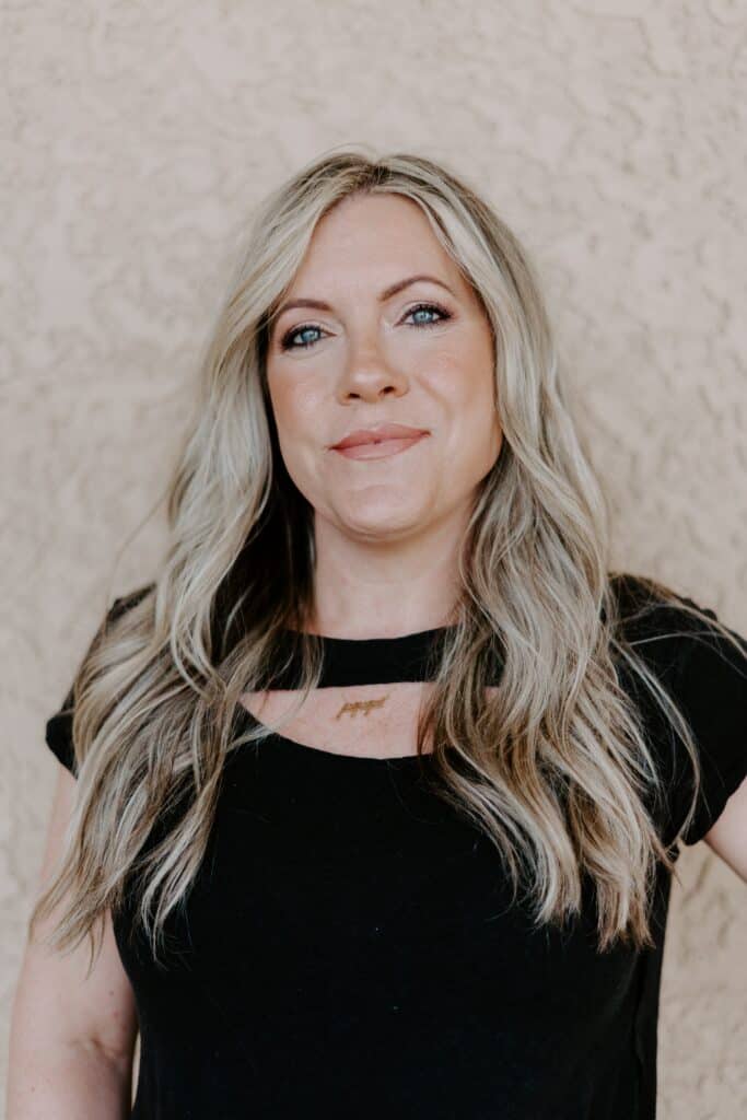 A woman with long, wavy blonde hair wearing a black top stands in front of a textured beige wall, looking at the camera.
