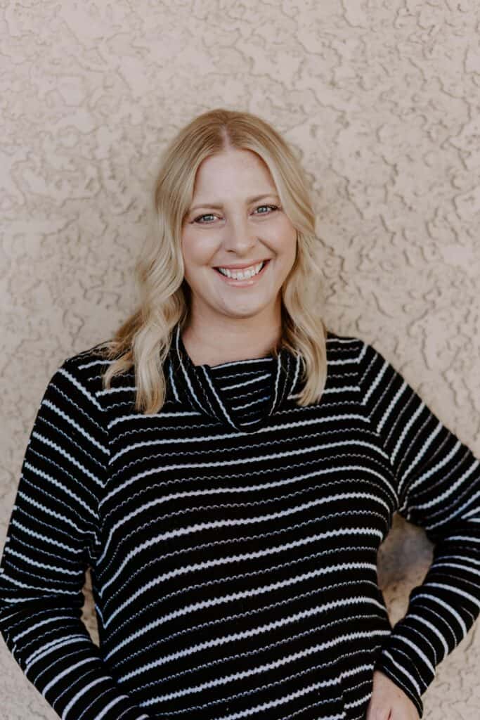 A woman with blonde hair wearing a black and white striped sweater stands in front of a textured beige wall, smiling at the camera.