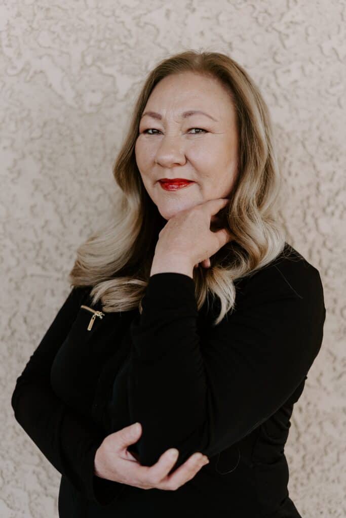 A woman with long, blonde hair and red lipstick poses with one hand under her chin, standing against a textured light-colored wall.