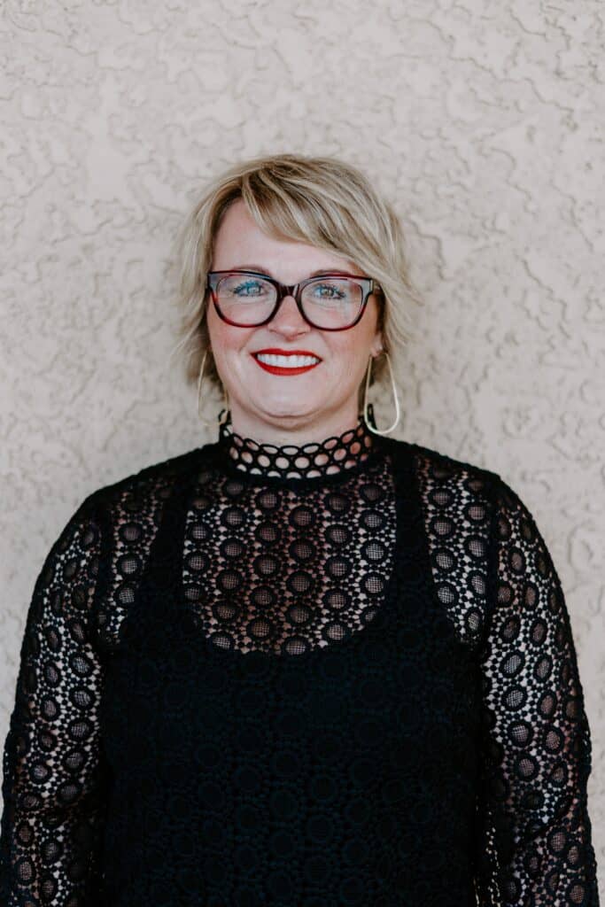A woman with short blonde hair, glasses, and red lipstick wears a black lace top and stands in front of a beige textured wall, smiling at the camera.