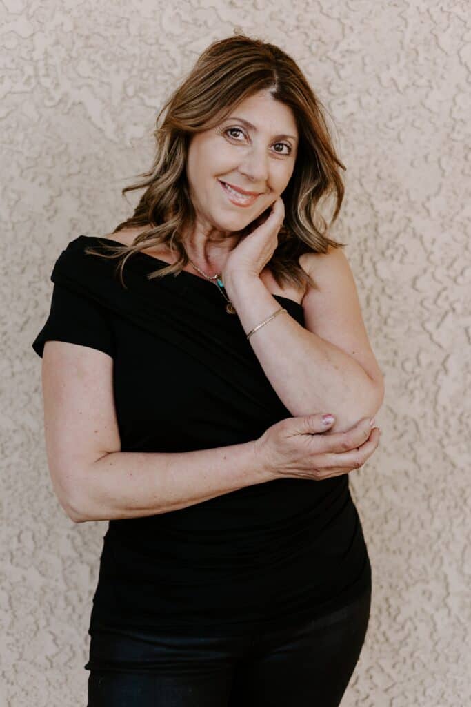 A woman with wavy brown hair wearing a black top stands in front of a textured beige wall, smiling with one hand touching her face and the other holding her elbow.