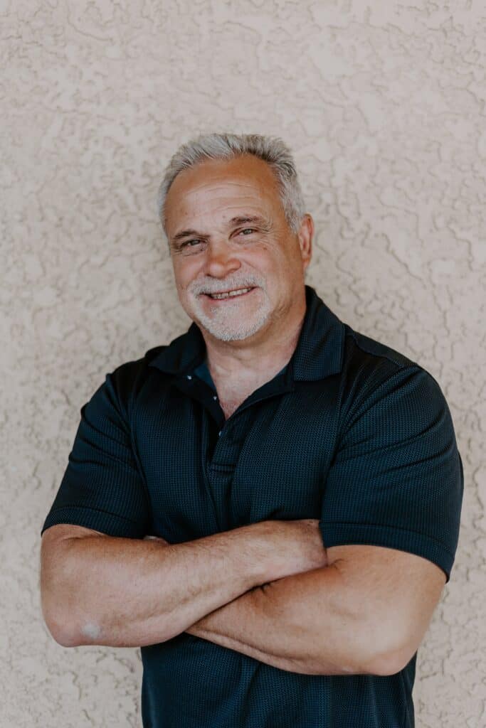 Older man with gray hair and a short beard, wearing a black polo shirt, standing with arms crossed and smiling in front of a textured beige wall.