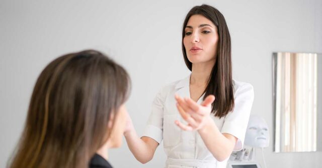 A woman in a white coat discusses lip filler options with another woman in an office setting, gesturing with her hands. A mask-like device is visible in the background.