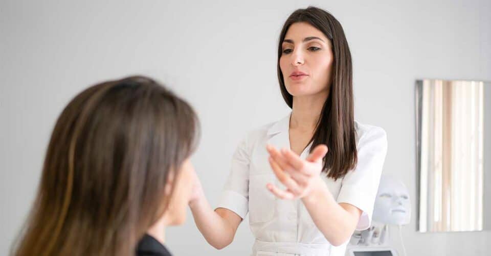 A woman in a white coat discusses lip filler options with another woman in an office setting, gesturing with her hands. A mask-like device is visible in the background.
