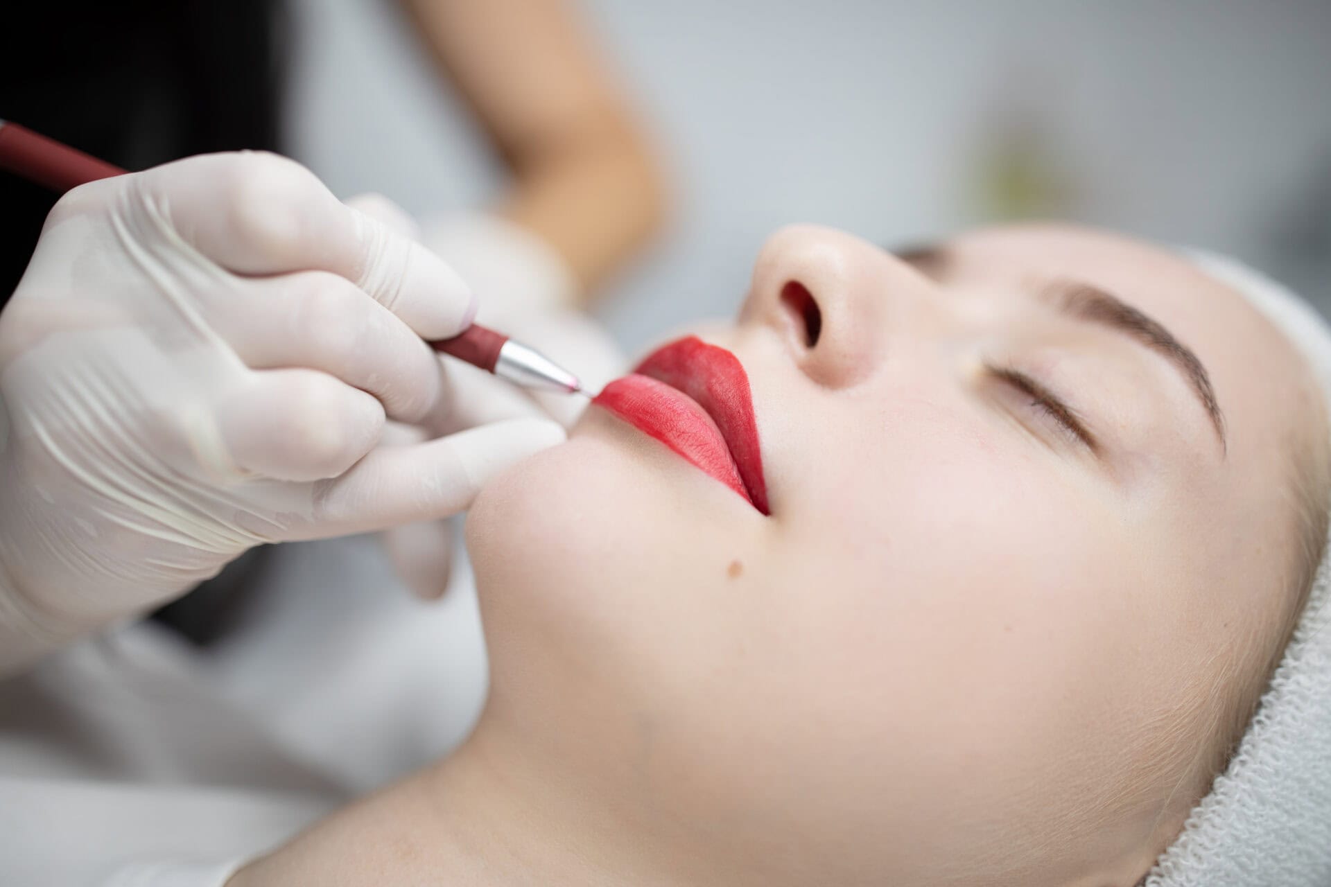Close-up of a person with closed eyes receiving a cosmetic lip tattoo procedure, while a gloved hand applies pigment to their lips—a great alternative to lip fillers from your local provider.