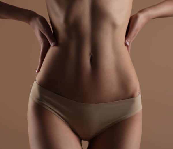 A woman wearing beige underwear stands with hands on her hips, showing her bare torso and abdomen against a plain brown background.