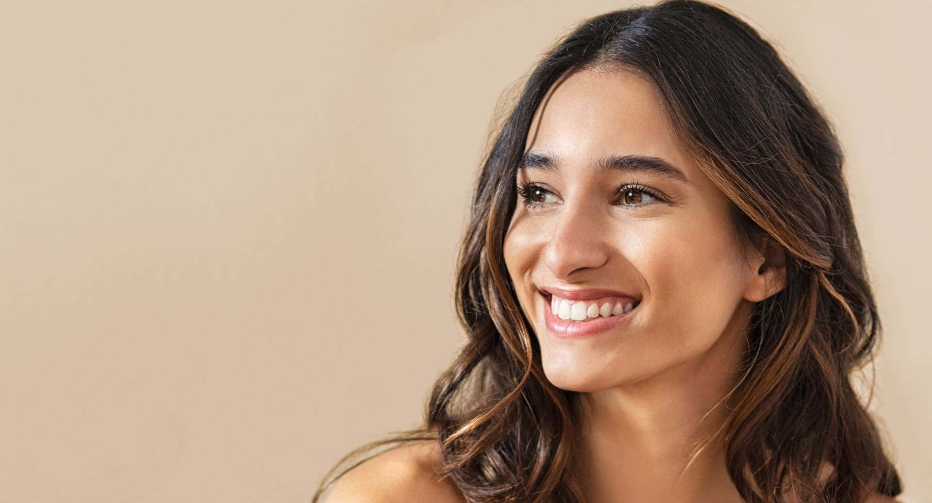 Woman with long dark hair smiling and looking to the right, set against a plain beige background.