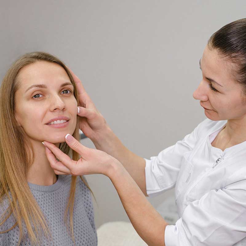 A woman in a gray sweater sits while a medical professional in a white coat examines her face, preparing for a Botox cosmetic treatment in a clinical setting.