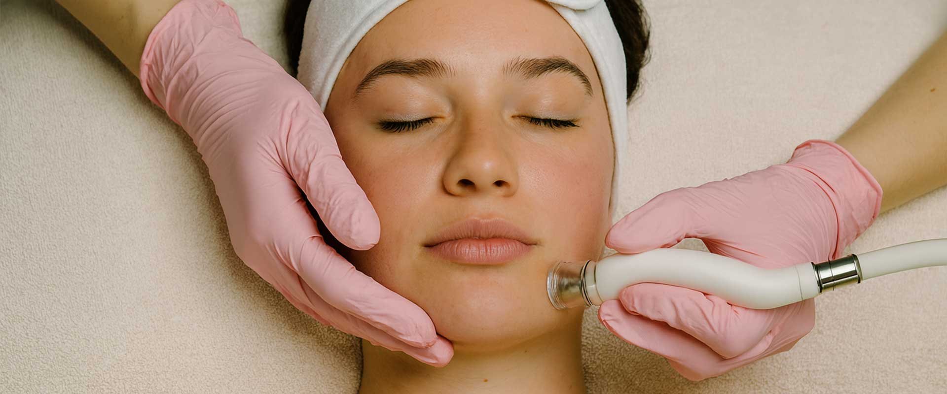 A person receives a facial treatment with a skincare device while wearing a headband, with a practitioner’s gloved hands holding the device and supporting the chin.