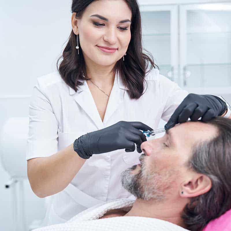 A woman in medical attire and black gloves administers an injection to a man's forehead in a clinical setting.