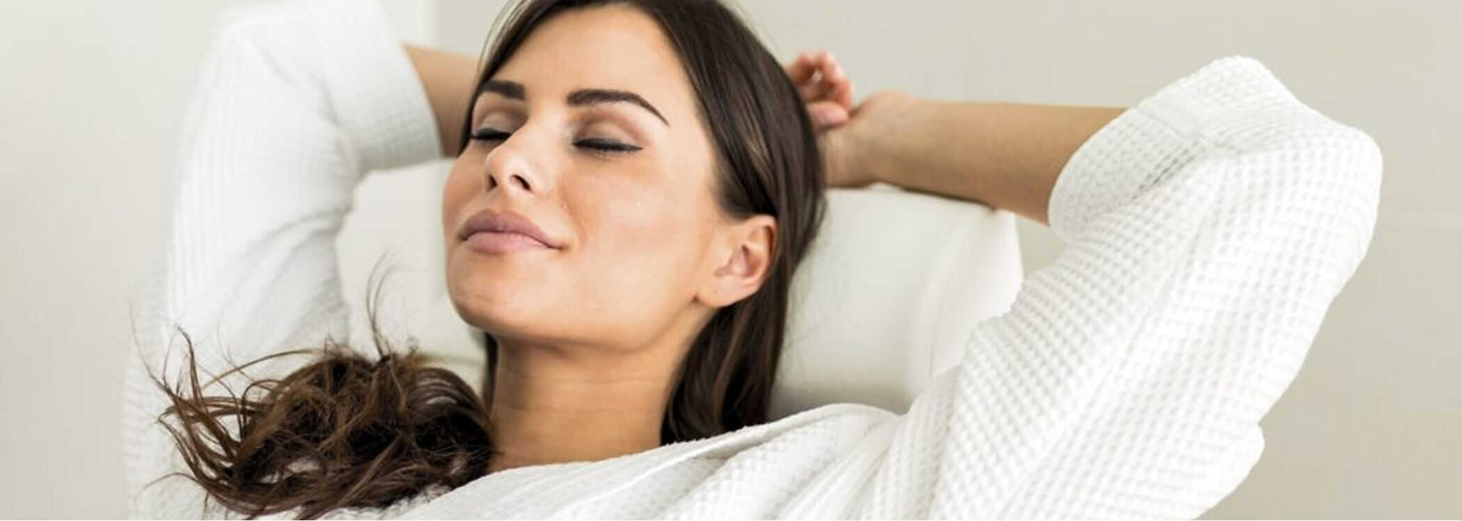 A woman in a white bathrobe leans back with her arms behind her head and eyes closed, appearing relaxed.