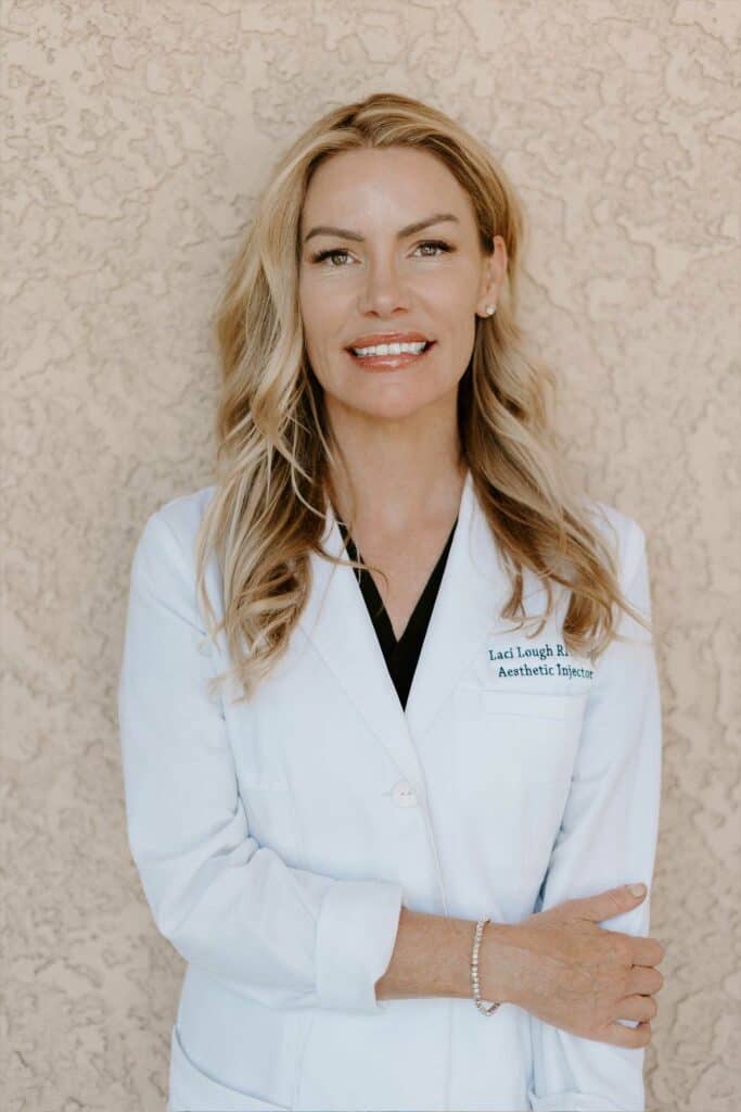 A woman with long blonde hair, wearing a white medical coat, stands in front of a textured beige wall, smiling with her arms gently crossed.