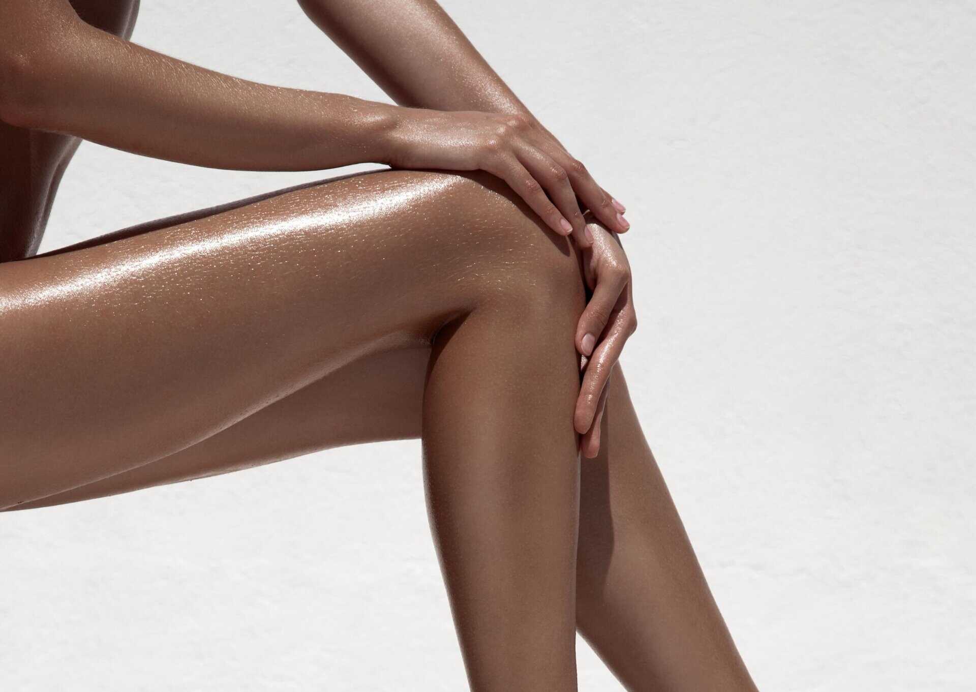 A close-up of a person's hands resting on their shiny, moisturized leg against a plain white background.