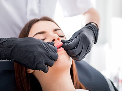 A person wearing black gloves massages the lips and face of a woman with closed eyes, who is seated in a chair, likely in a professional or medical setting.