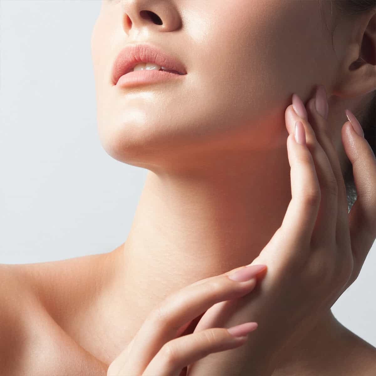 Close-up of a woman's lower face, neck, and hand with smooth skin and nude-colored lips and nails, against a light background.