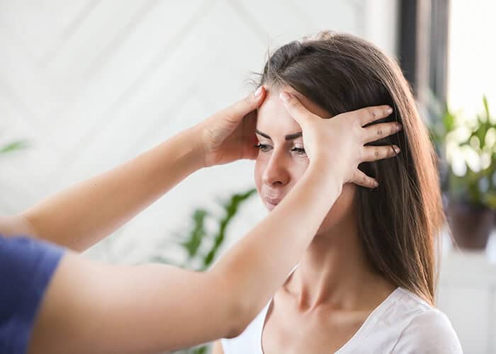 A person gently places their hands on another person’s forehead and temples, possibly helping to relieve headaches or migraines, or conducting a relaxation technique indoors.