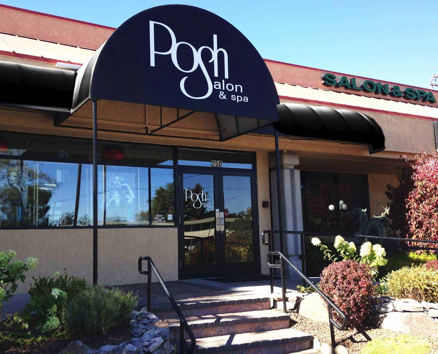 Exterior view of Posh Salon & Spa with black awning, glass entrance door, and steps leading up to the entrance, surrounded by landscaping and clear blue sky.