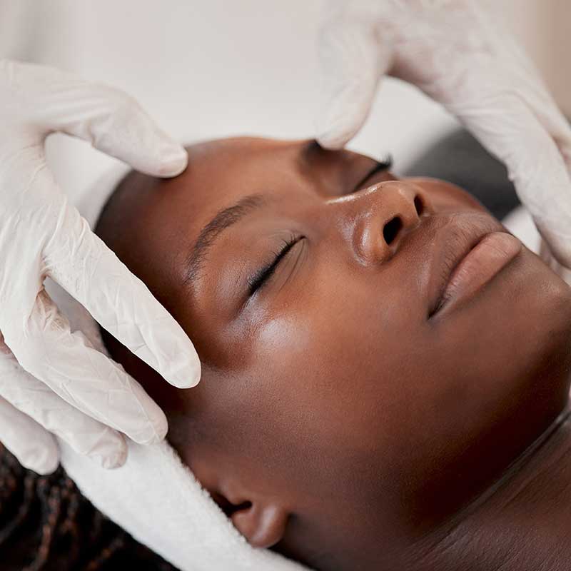 A person with closed eyes receives a facial massage from someone wearing white gloves. The person has a towel wrapped around their head.