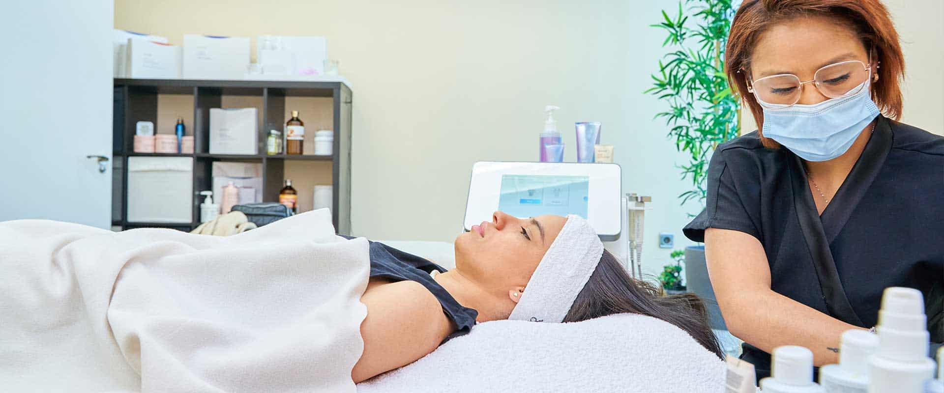 A woman lies on a treatment bed with a headband while a masked skincare professional prepares products in a clinic room.