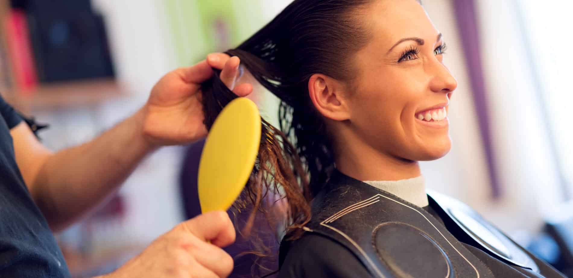 A hairdresser uses a yellow brush to comb a smiling woman's wet hair at a salon.