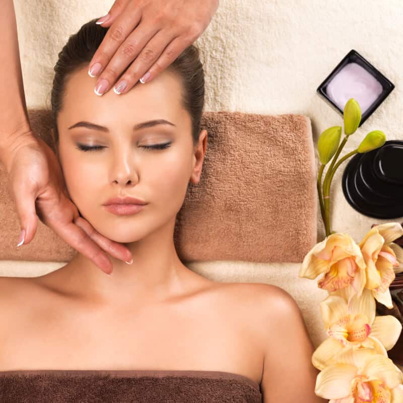 A woman lies on a towel with her eyes closed, receiving a facial massage, with flowers and skincare products arranged nearby.