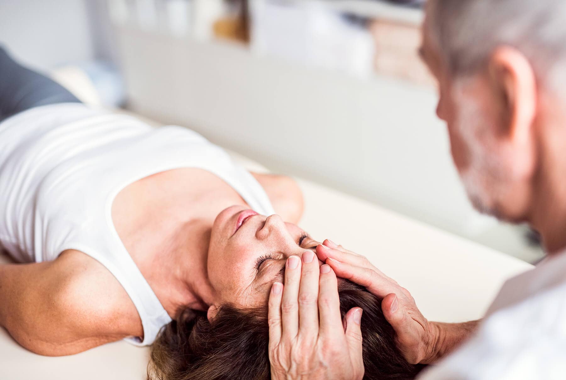 A woman lies on her back while a person places their hands on her forehead, appearing to perform a therapeutic technique often used for relieving headaches or migraines.