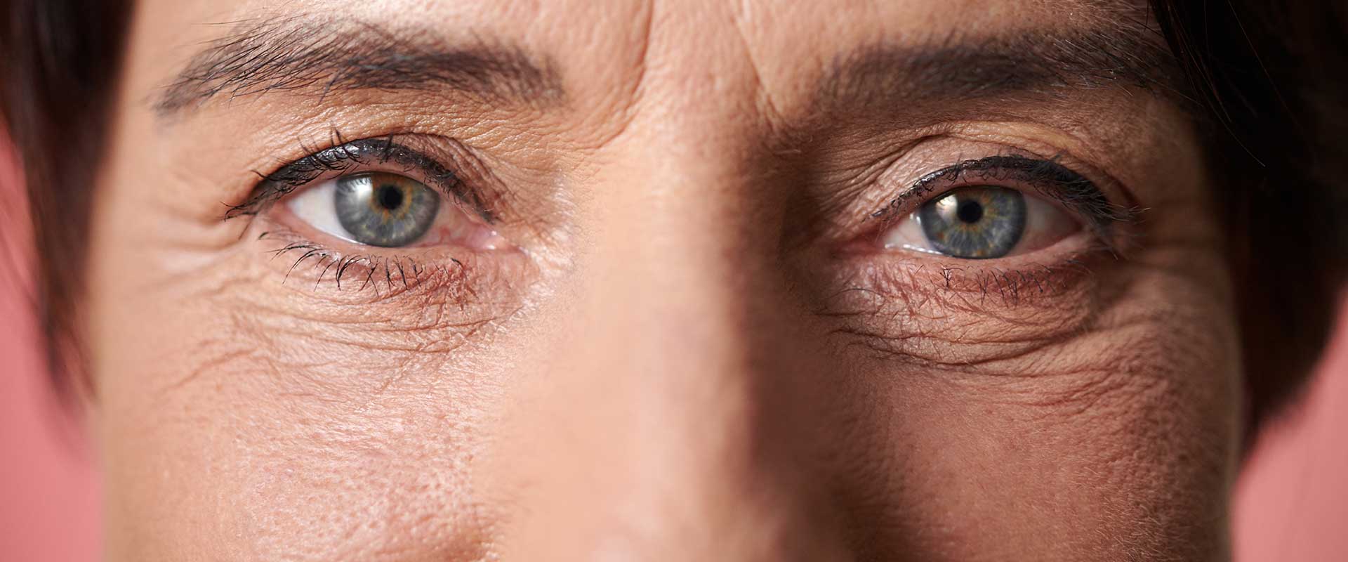 Close-up of a person's face focusing on their blue eyes and the surrounding wrinkles, highlighting under eye issues against a pink background.