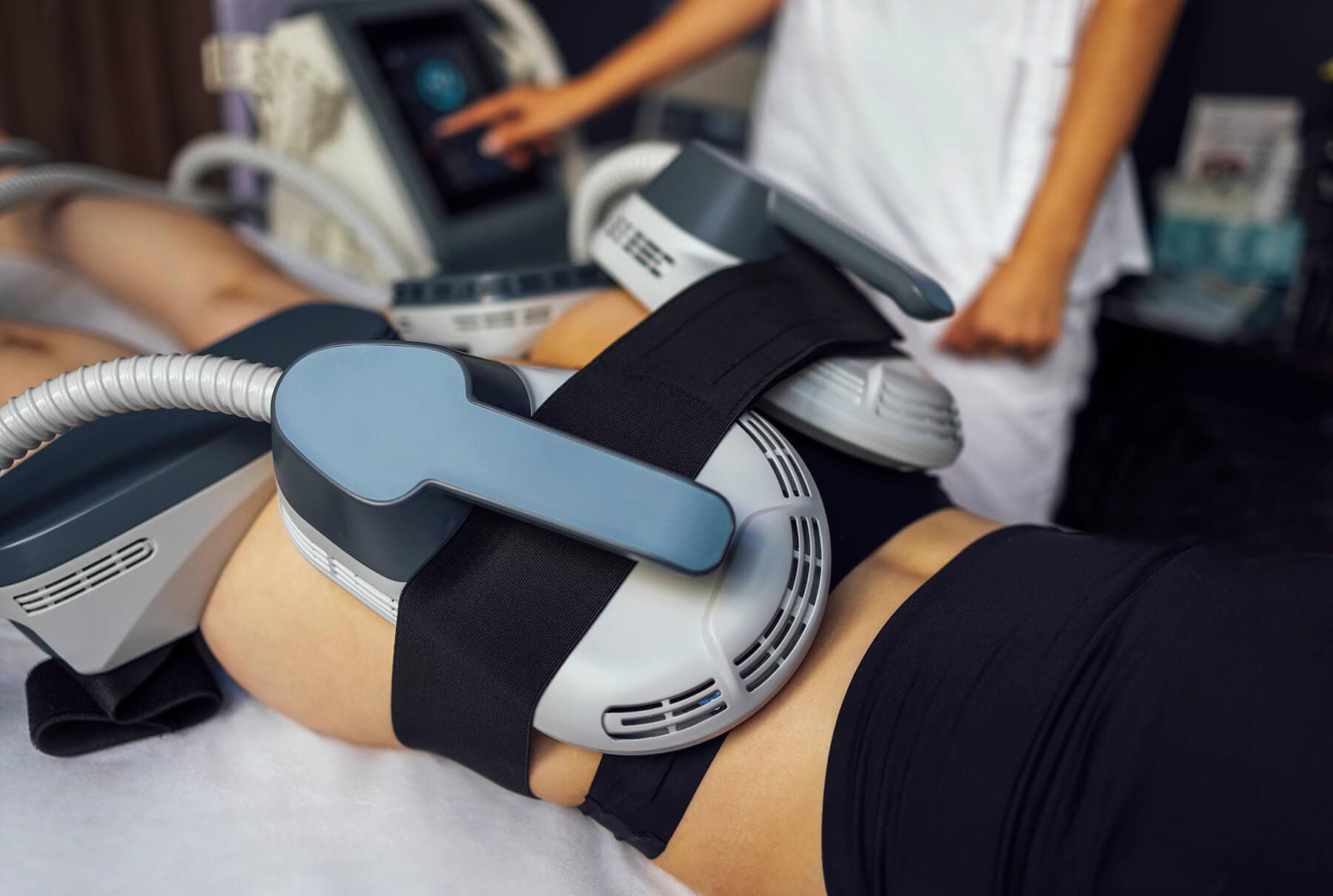 Person undergoing a non-invasive body sculpting treatment with large applicator devices, while a clinician operates the nearby Emsculpt Neo machine.