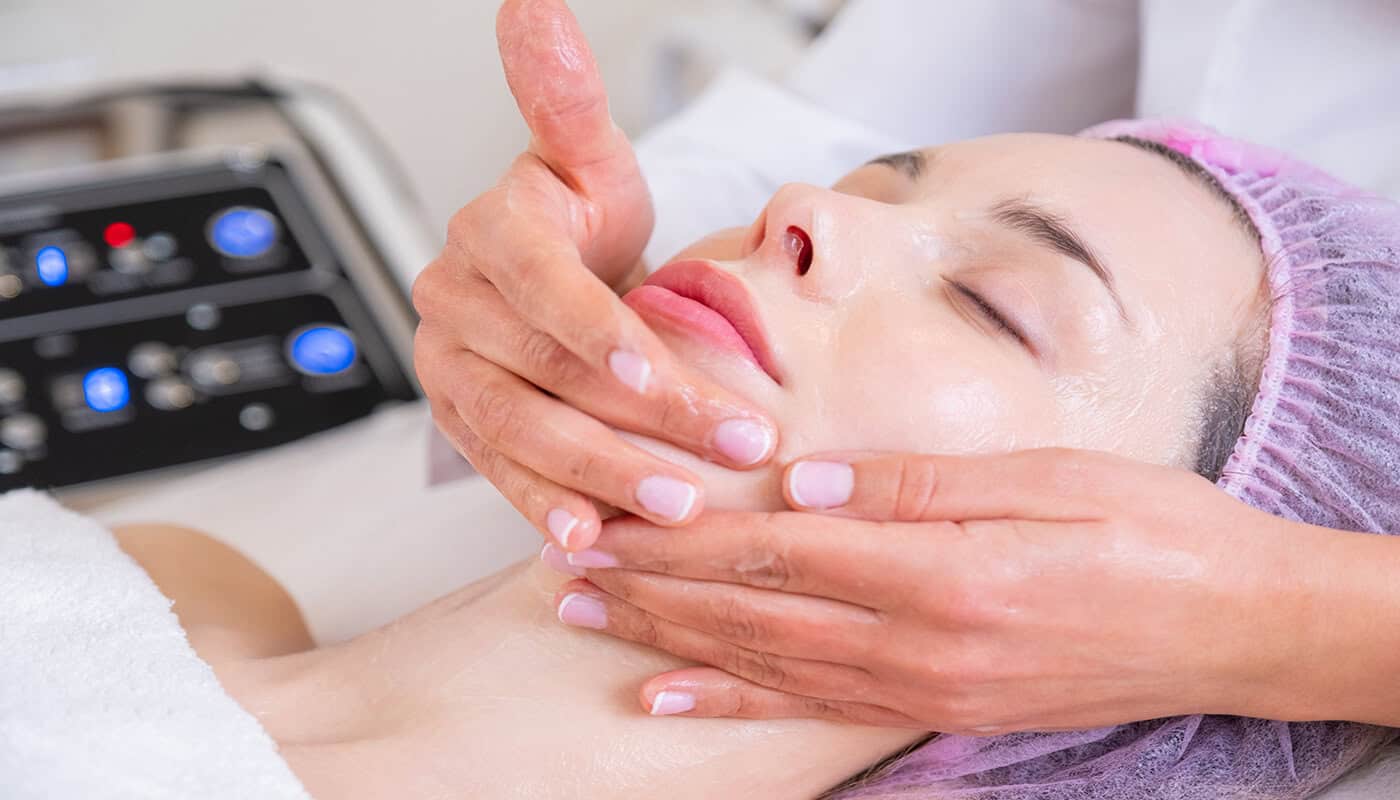 Person receiving a facial treatment at a spa, eyes closed and wearing a hair cover, while a professional gently massages their face to enhance the results of glow treatments.